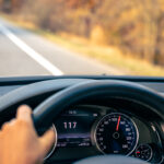 Volant d'une voiture en circulation avec une main dessus et la vitesse de 117 affichée sur le tableau de bord